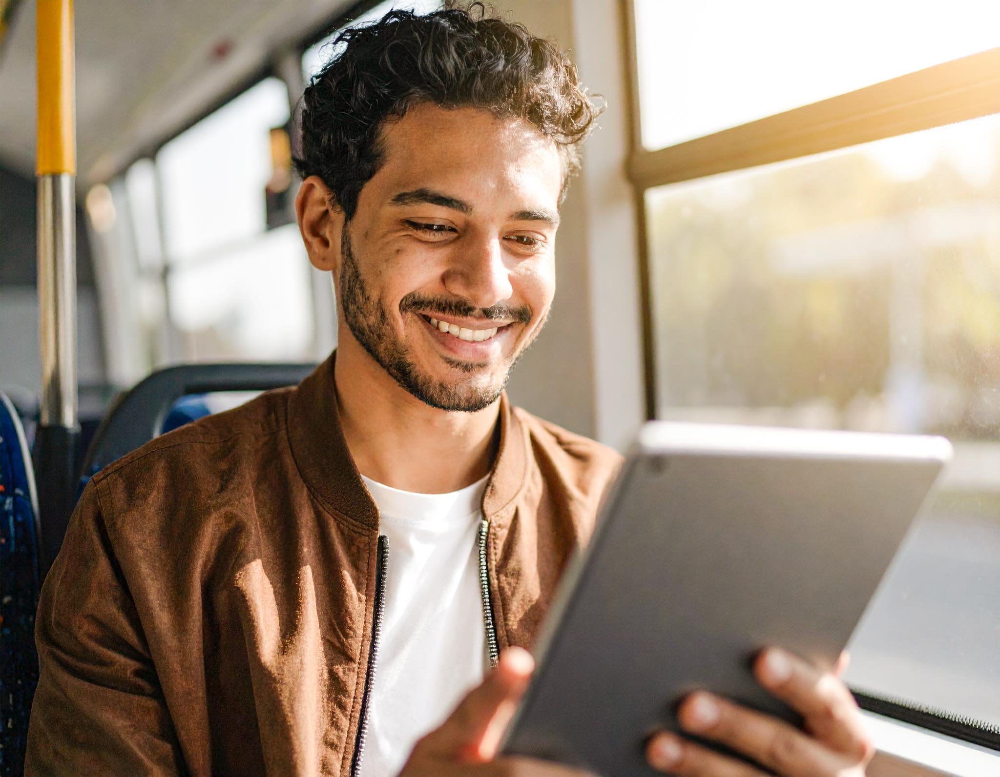 Eine junger Mann sitzt an einem sonnigen Tag in einer Straßenbahn. Er liest etwas auf seinem-2 Junger Mann mit Bart liest auf einem Tablet im Zug – steht für flexiblen digitalen Zugang zur Zeitung unterwegs