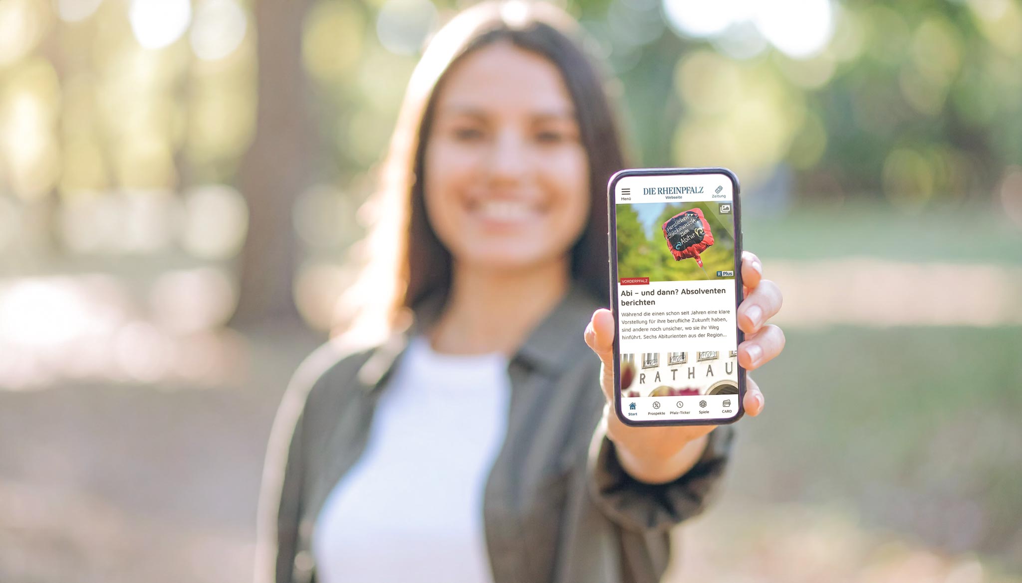 Frau mit Handy vorne Bearbeitet-2 Junge Frau hält lächelnd ein Smartphone mit der geöffneten RHEINPFALZ-App in die Kamera – Symbol für mobile Nutzung und digitalen Zugang