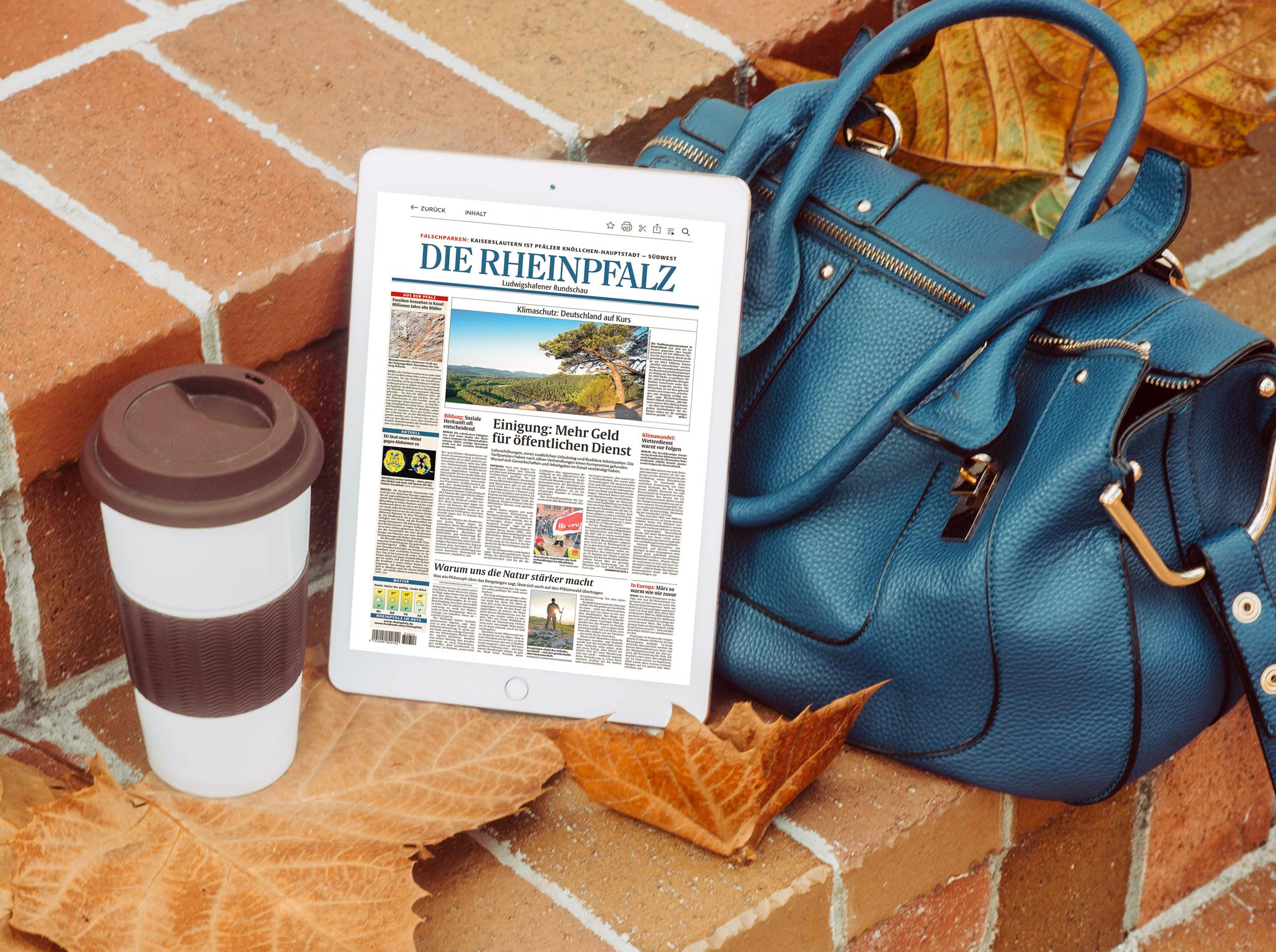 Ein Tablet auf einer Backsteintreppe zeigt das E-Paper der Zeitung DIE RHEINPFALZ. Daneben liegt eine blaue Handtasche, ein weiß-brauner Mehrwegbecher und einige herbstliche Blätter.