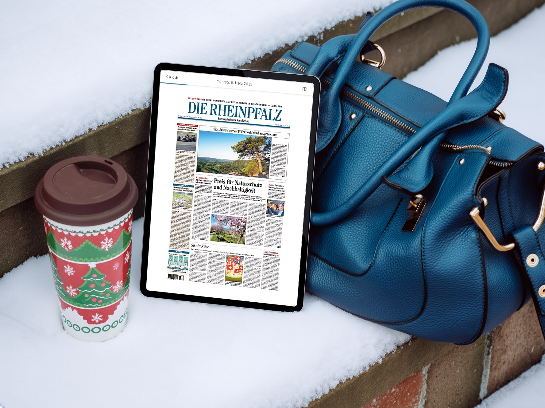 Ein Tablet auf einer verschneiten Backsteintreppe zeigt das E-Paper der Zeitung DIE RHEINPFALZ. Rechts neben dem Tablet liegt eine blaue Handtasche auf der schneebedeckten Stufe, links neben dem Tablet steht ein Mehrwegbecher mit Weihnachtsmotiv.