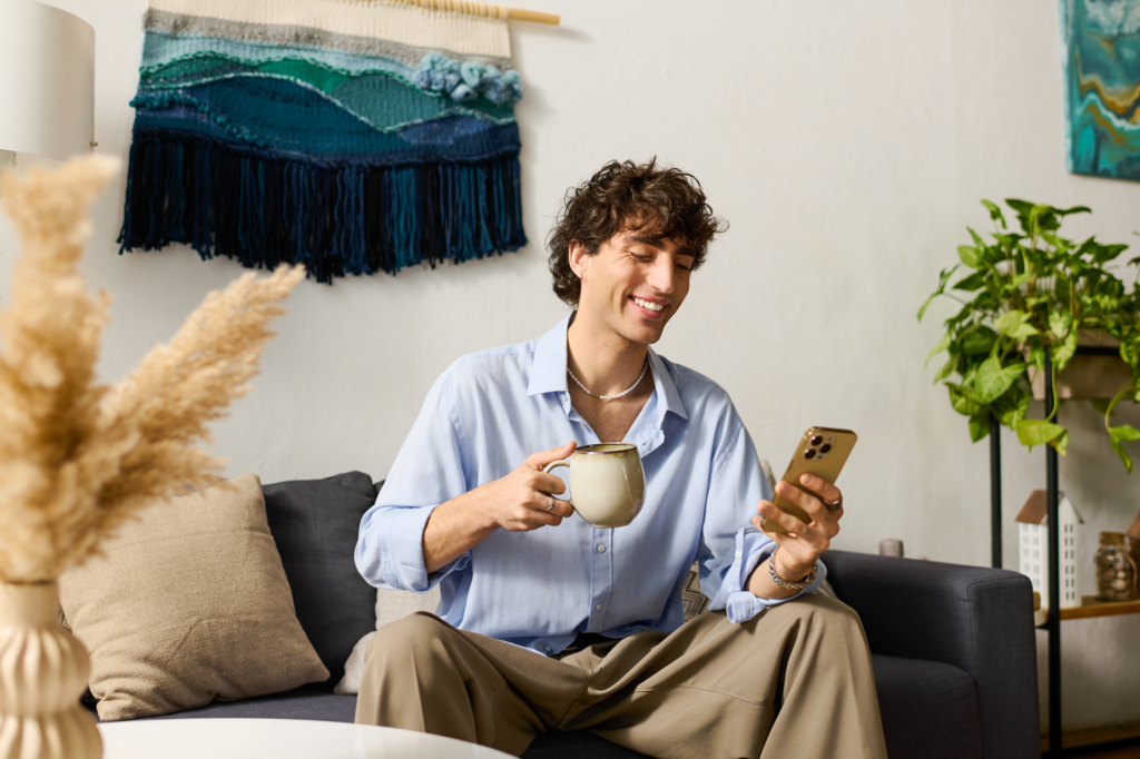 Junger Mann sitzt lächelnd auf dem Sofa und liest die RHEINPFALZ auf seinem Smartphone.
