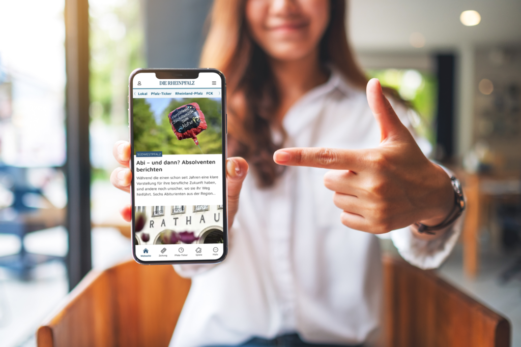 Eine junge Frau zeigt auf ihr Smartphone, dessen Display sie in den Fokus der Kamera hält. Auf dem Smartphone ist die Startseite der RHEINPFALZ-App zu sehen.
